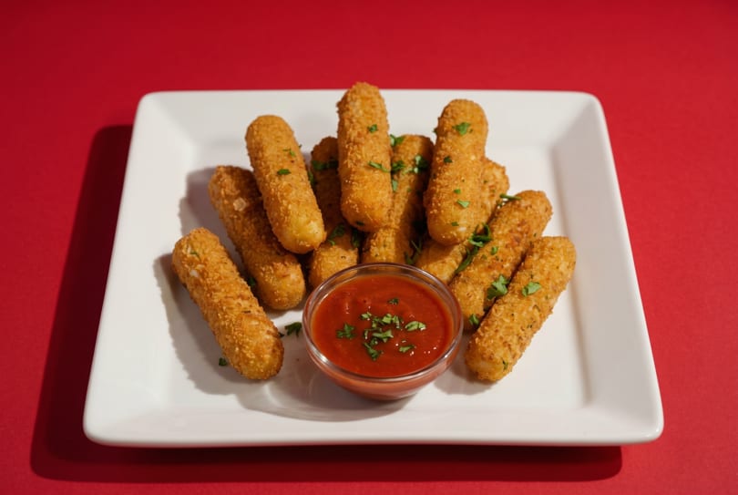 Golden-brown mozzarella sticks with parsley flakes, served with a side of marinara sauce on a white plate.