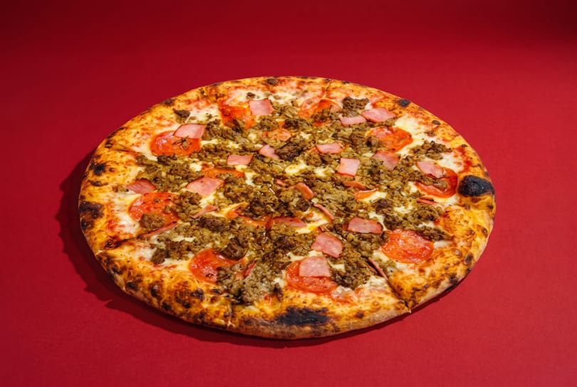 A whole meat lover's pizza with pepperoni, sausage, and ham on a red background.