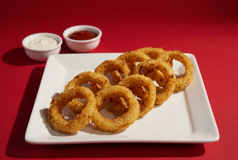 Golden-brown onion rings on a white rectangular plate with two dipping sauces on a red background.
