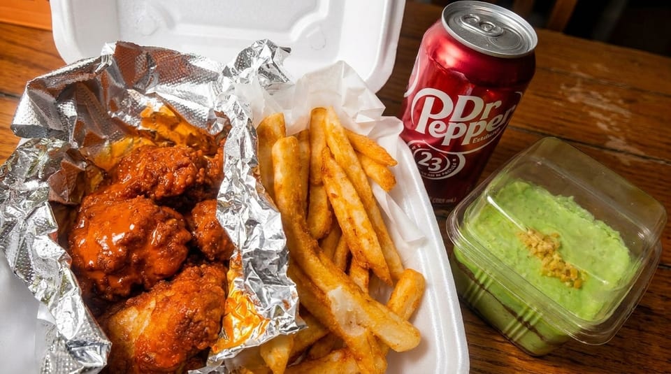 Spicy chicken wings, french fries, guacamole, and a Dr. Pepper in a takeout container.