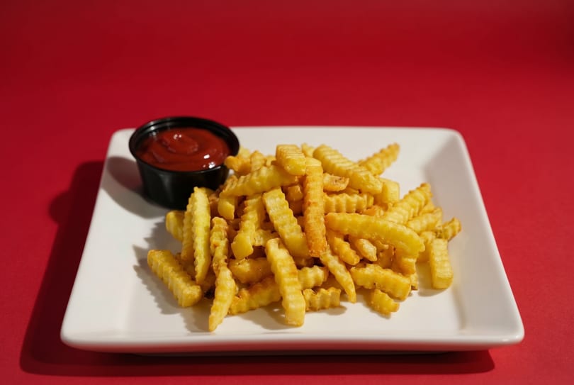 A white rectangular plate holds a pile of golden crinkle-cut fries and a small cup of ketchup.