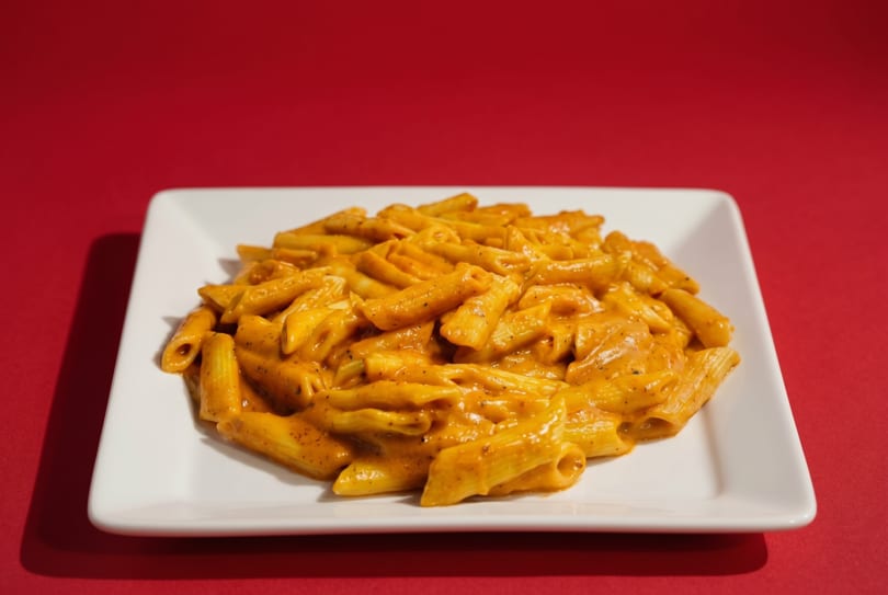 Penne pasta coated in a creamy orange sauce, served on a white rectangular plate against a red background.