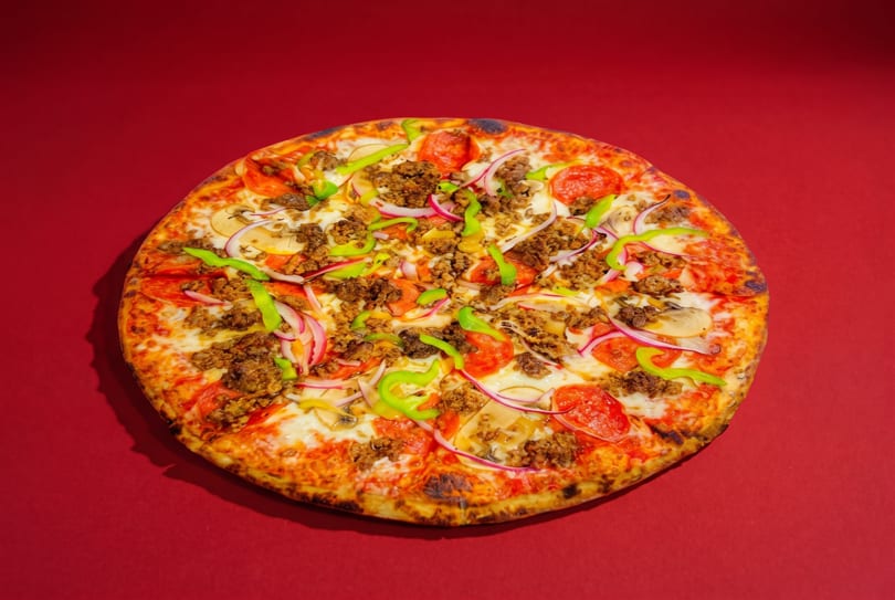A whole pizza with pepperoni, ground meat, green peppers, red onions, and mushrooms on a red background.