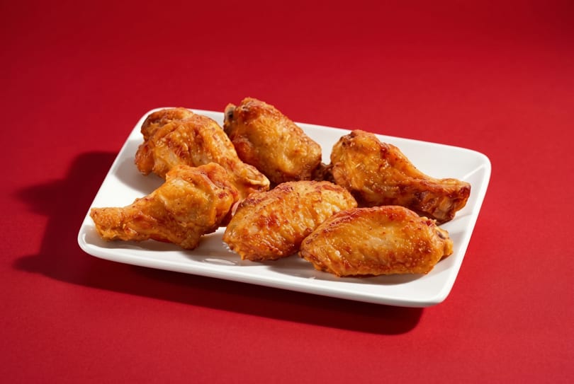 Six golden-brown fried chicken wings are arranged on a white rectangular plate against a red background.